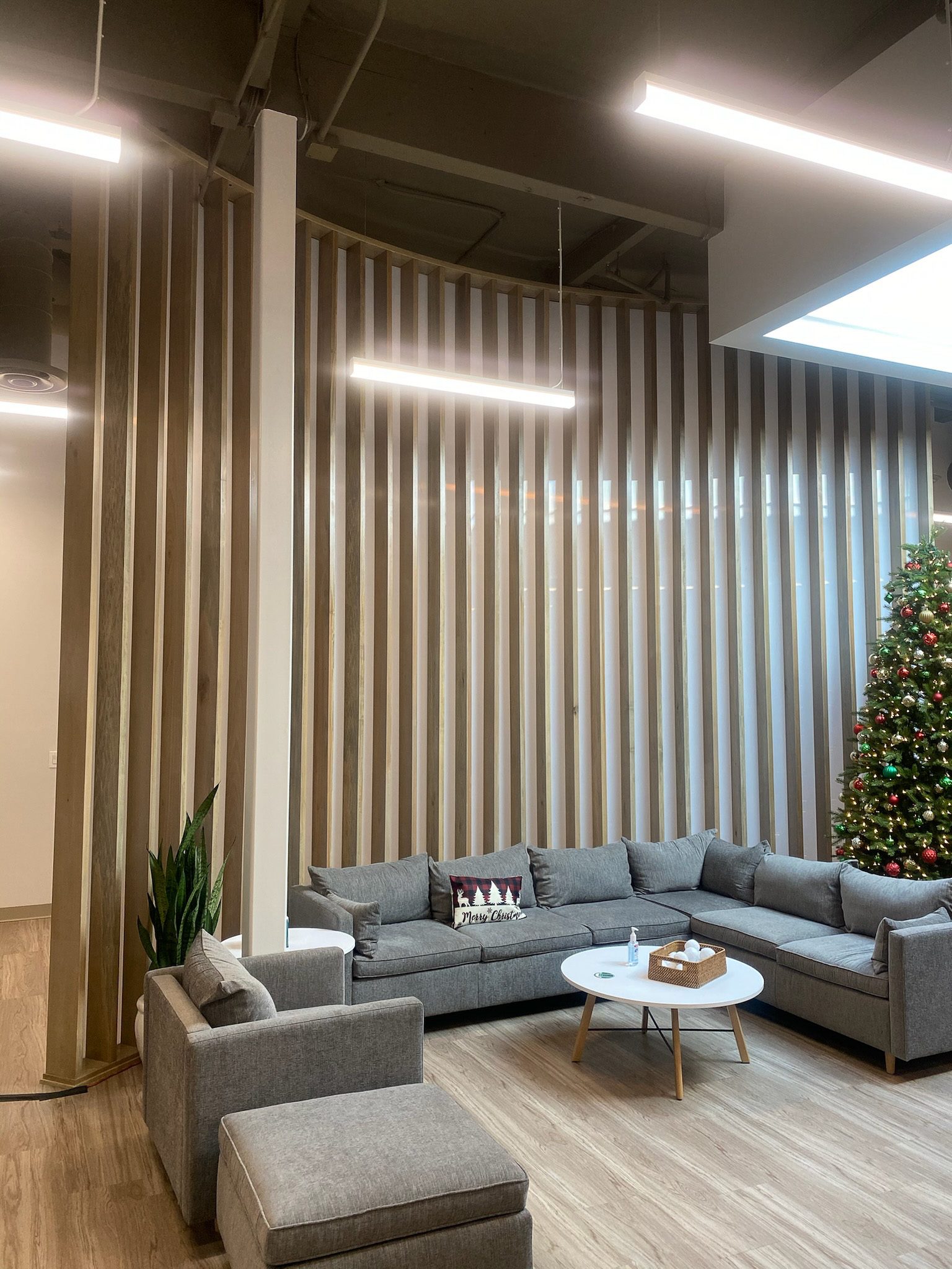 Modern office lounge area with a gray sectional sofa, wooden slatted walls, and hanging linear lights. a christmas tree is visible in the background.